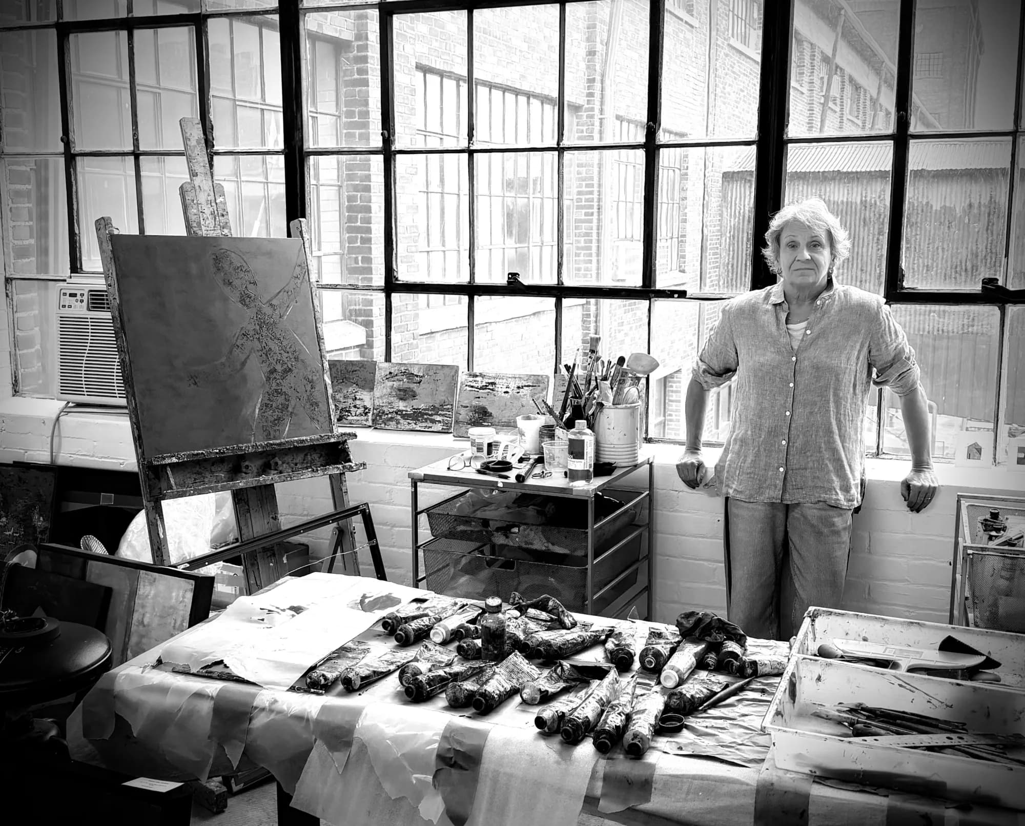 Kate Singer standing by the window in her studio, beside an easel and a table of oil paint tubes
